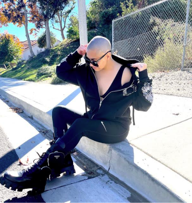 Sabryna Rocha sitting on a curb all in black streetwear and sunglasses with shaved head looking very chic