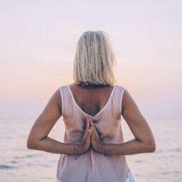 A blond woman with a white tank top faces the ocean with her hands in mudra behind her back